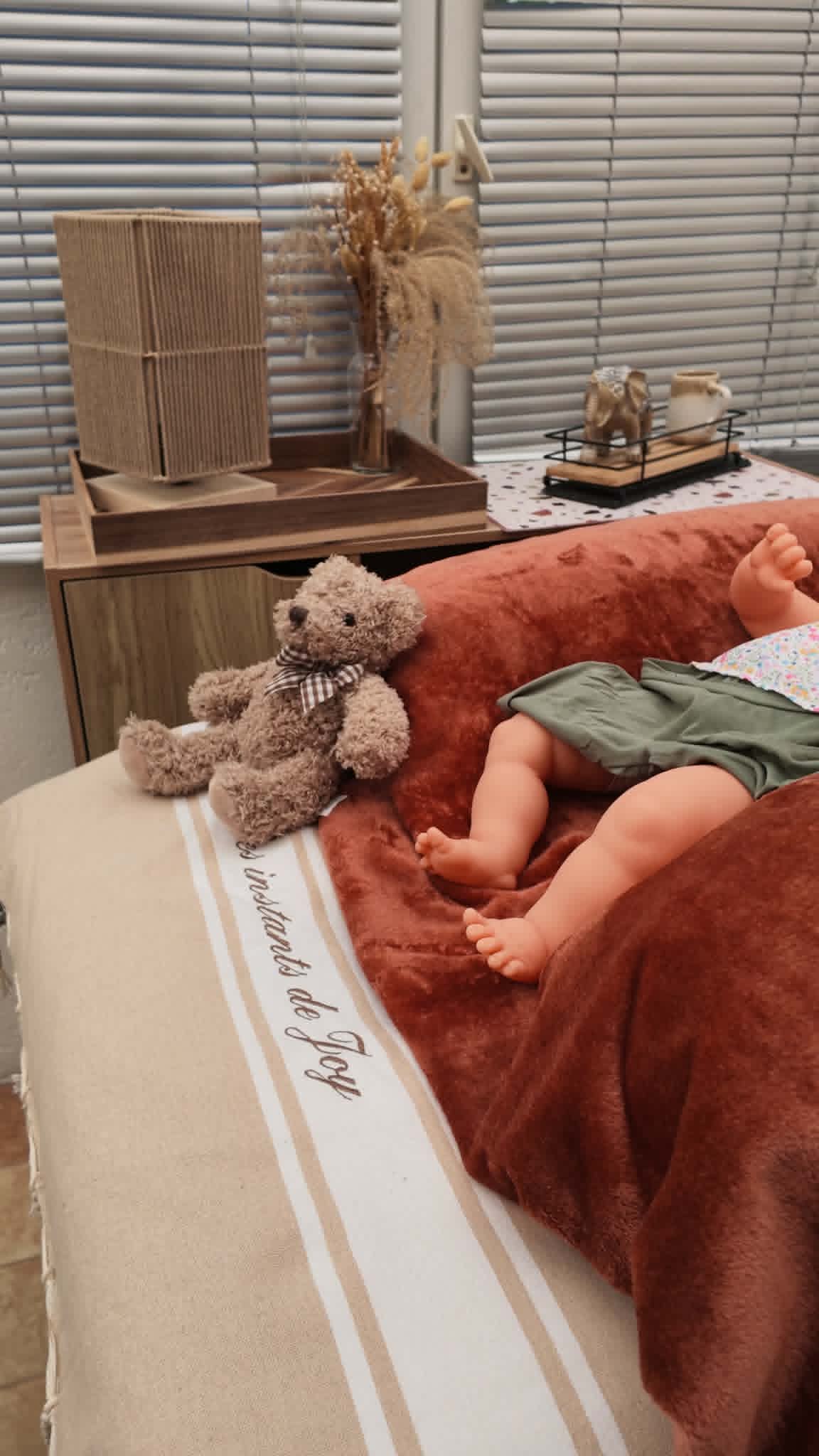 Poupon installé sur un plaid pour l’atelier massage bébé, à côté d’un ours en peluche, chez une famille près de Nantes.
